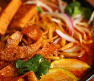Bowl of vegan khao soi containing seared tofu, baked dumpling wrapper strips, cilantro, and lemon, in a coconut-curry broth