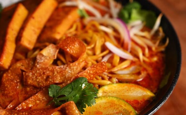Bowl of vegan khao soi containing seared tofu, baked dumpling wrapper strips, cilantro, and lemon, in a coconut-curry broth
