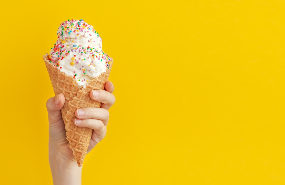Child’s hand holding vanilla ice cream cone with sprinkles against a yellow background, to illustrate ingredients connected to fossil fuels in food