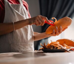 Young woman wearing an apron peeling a sweet potato in a kitchen, to illustrate an article about the anti-cancer benefits of sweet potatoes