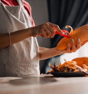 Young woman wearing an apron peeling a sweet potato in a kitchen, to illustrate an article about the anti-cancer benefits of sweet potatoes