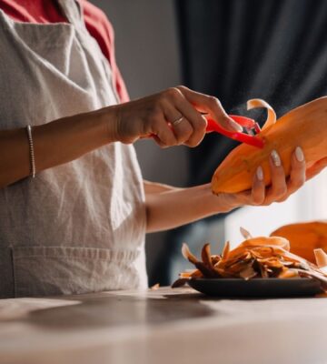 Young woman wearing an apron peeling a sweet potato in a kitchen, to illustrate an article about the anti-cancer benefits of sweet potatoes