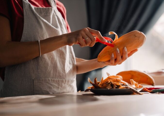 Young woman wearing an apron peeling a sweet potato in a kitchen, to illustrate an article about the anti-cancer benefits of sweet potatoes