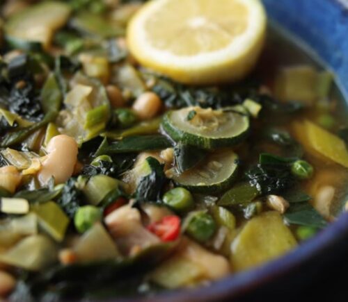 Vegan longevity stew with cannellini beans and leafy greens, served in a blue bowl and topped with a decorative lemon slice