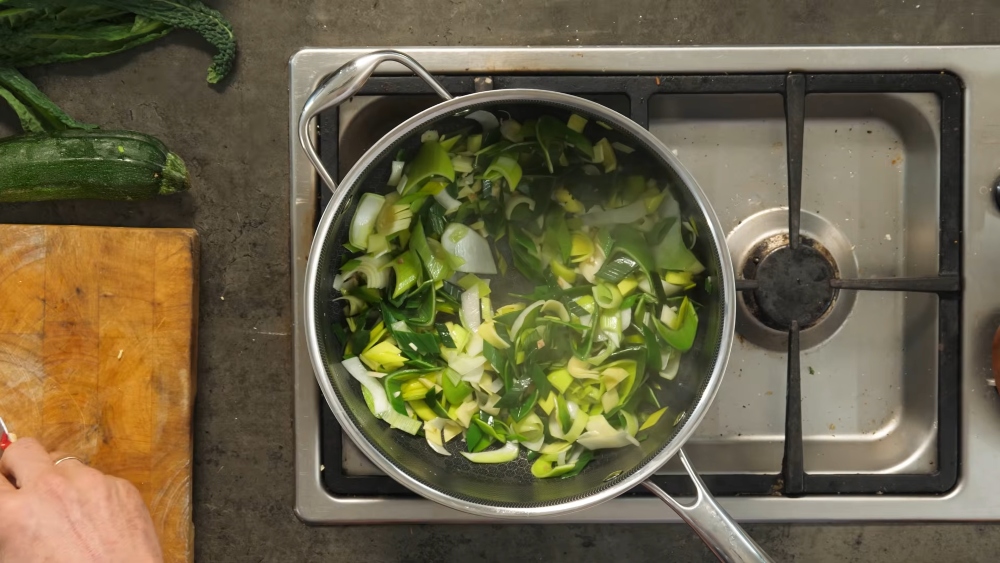Shot of the greens of this vegan longevity stew cooking on the stove as the twin chops more ingredients on a cutting board