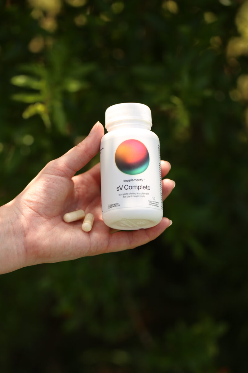 Photo shows someone's hand as they hold up a bottle and two capsules of supplementV's sV Complete, a vegan all-in-one supplement that the company says is more than just a multivitamin