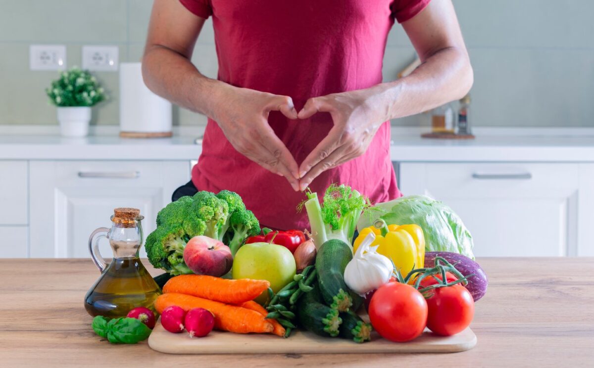 Photo shows an athletic-looking man making a heart shape with his hands above a large pile of plant-based foods, including fruit, vegetables, and olive oil. The American Heart Association (AHA) recently updated its guidelines for cardiovascular health and recommends that people eat more plant protein and less meat