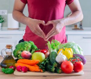 Photo shows an athletic-looking man making a heart shape with his hands above a large pile of plant-based foods, including fruit, vegetables, and olive oil. The American Heart Association (AHA) recently updated its guidelines for cardiovascular health and recommends that people eat more plant protein and less meat