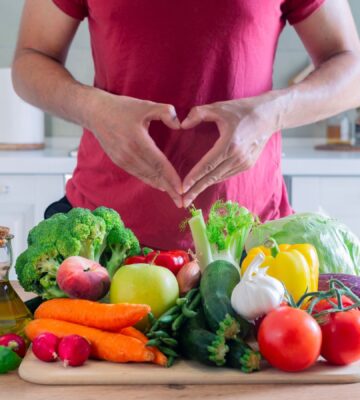 Photo shows an athletic-looking man making a heart shape with his hands above a large pile of plant-based foods, including fruit, vegetables, and olive oil. The American Heart Association (AHA) recently updated its guidelines for cardiovascular health and recommends that people eat more plant protein and less meat