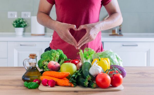 Photo shows an athletic-looking man making a heart shape with his hands above a large pile of plant-based foods, including fruit, vegetables, and olive oil. The American Heart Association (AHA) recently updated its guidelines for cardiovascular health and recommends that people eat more plant protein and less meat