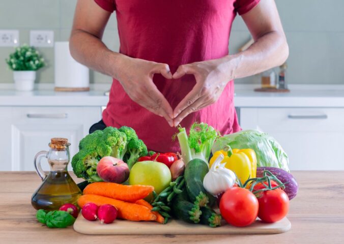 Photo shows an athletic-looking man making a heart shape with his hands above a large pile of plant-based foods, including fruit, vegetables, and olive oil. The American Heart Association (AHA) recently updated its guidelines for cardiovascular health and recommends that people eat more plant protein and less meat