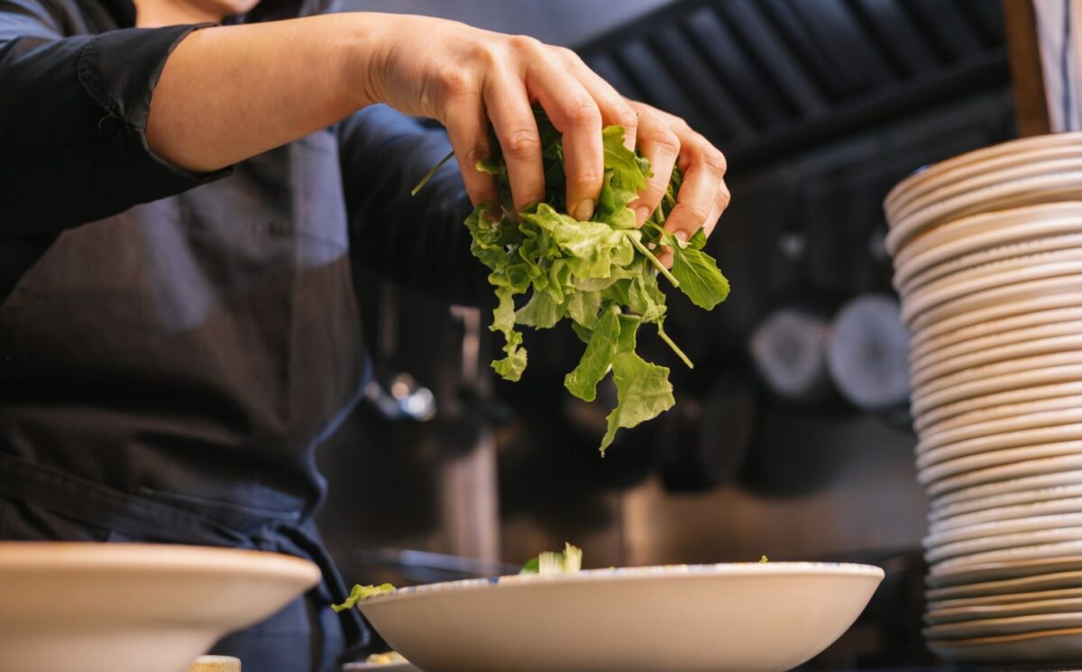 Photo shows someone in a dark uniform placing salad leaves into a bowl. UK prisons are now required to take imprisoned vegan people’s ethical beliefs into account while planning menus.