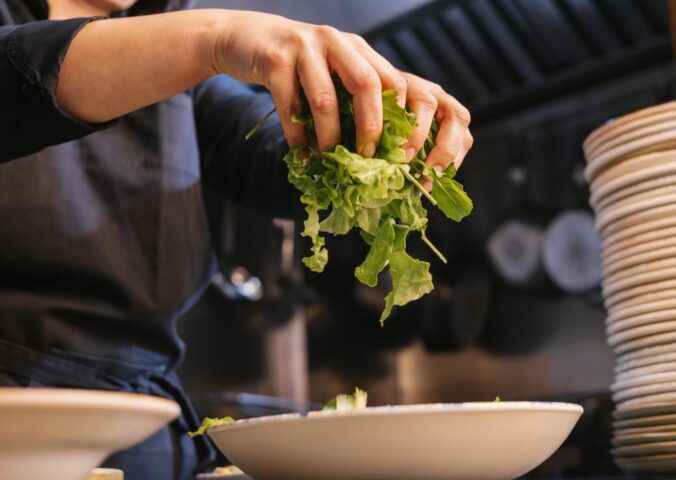 Photo shows someone in a dark uniform placing salad leaves into a bowl. UK prisons are now required to take imprisoned vegan people’s ethical beliefs into account while planning menus.