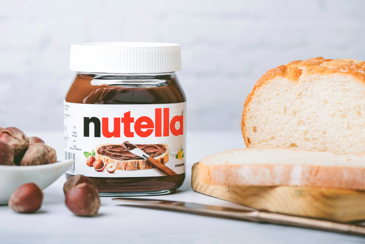 Photo shows a jar of classic Nutella chocolate spread next to a dish of hazelnuts and a sliced white loaf of bread. To celebrate World Nutella Day, Ferrero finally launched Nutella Peanut, which is both the first vegan Nutella product in North America and an entirely new flavor for the brand