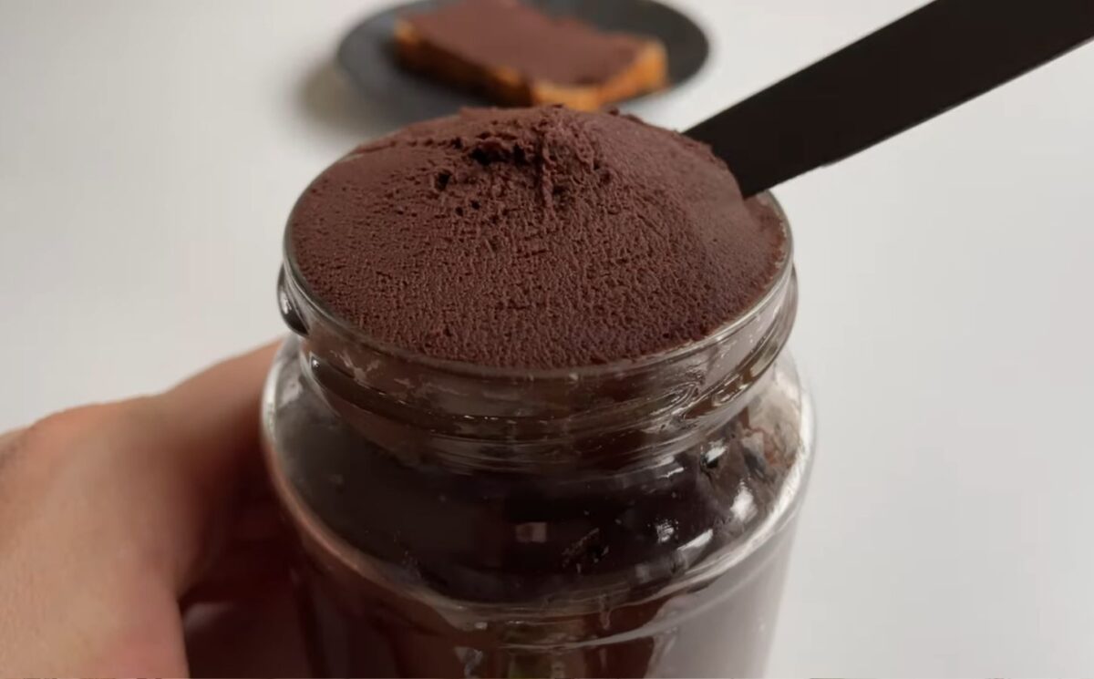 Glass jar of vegan chocolate chickpea spread with a slice of bread covered in the chocolate spread on a plate in the background