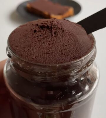 Glass jar of vegan chocolate chickpea spread with a slice of bread covered in the chocolate spread on a plate in the background