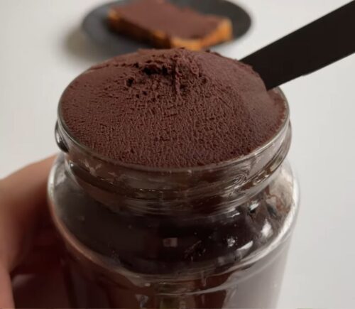 Glass jar of vegan chocolate chickpea spread with a slice of bread covered in the chocolate spread on a plate in the background