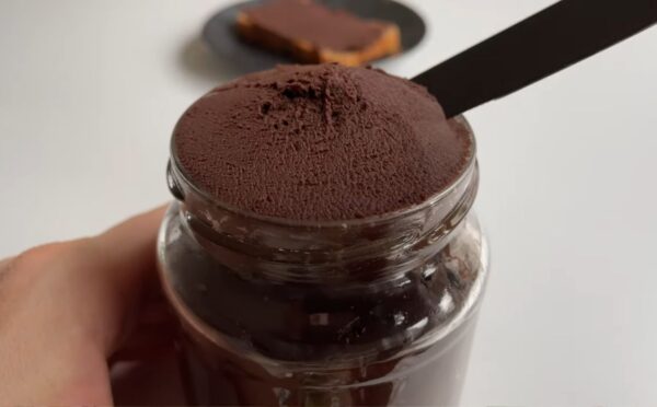 Glass jar of vegan chocolate chickpea spread with a slice of bread covered in the chocolate spread on a plate in the background