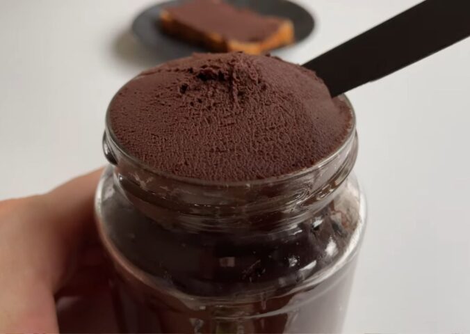 Glass jar of vegan chocolate chickpea spread with a slice of bread covered in the chocolate spread on a plate in the background