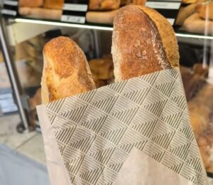 Two baguettes in a paper bag inside a Parisian bakery, with a display of baked goods in the background, illustrating how bread can contribute to daily vegan protein intake