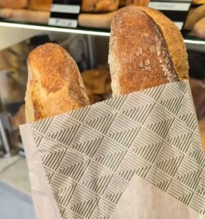 Two baguettes in a paper bag inside a Parisian bakery, with a display of baked goods in the background, illustrating how bread can contribute to daily vegan protein intake