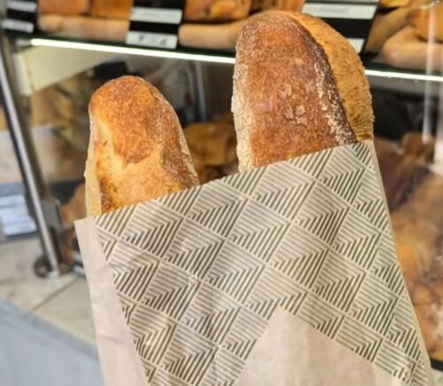 Two baguettes in a paper bag inside a Parisian bakery, with a display of baked goods in the background, illustrating how bread can contribute to daily vegan protein intake