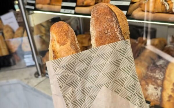 Two baguettes in a paper bag inside a Parisian bakery, with a display of baked goods in the background, illustrating how bread can contribute to daily vegan protein intake