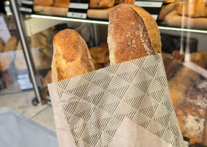Two baguettes in a paper bag inside a Parisian bakery, with a display of baked goods in the background, illustrating how bread can contribute to daily vegan protein intake