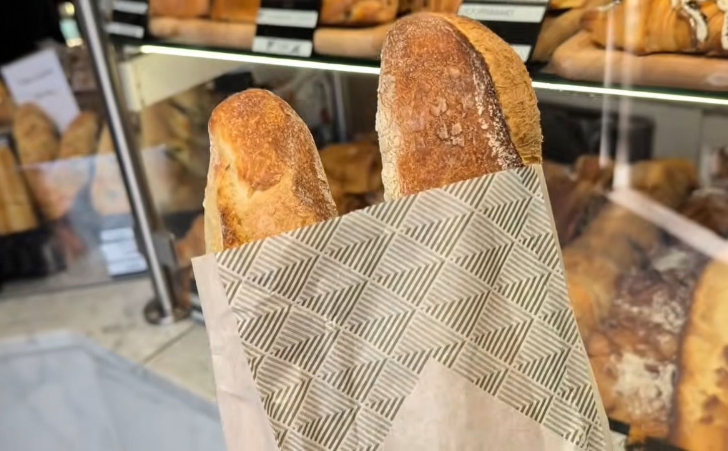 Two baguettes in a paper bag inside a Parisian bakery, with a display of baked goods in the background, illustrating how bread can contribute to daily vegan protein intake