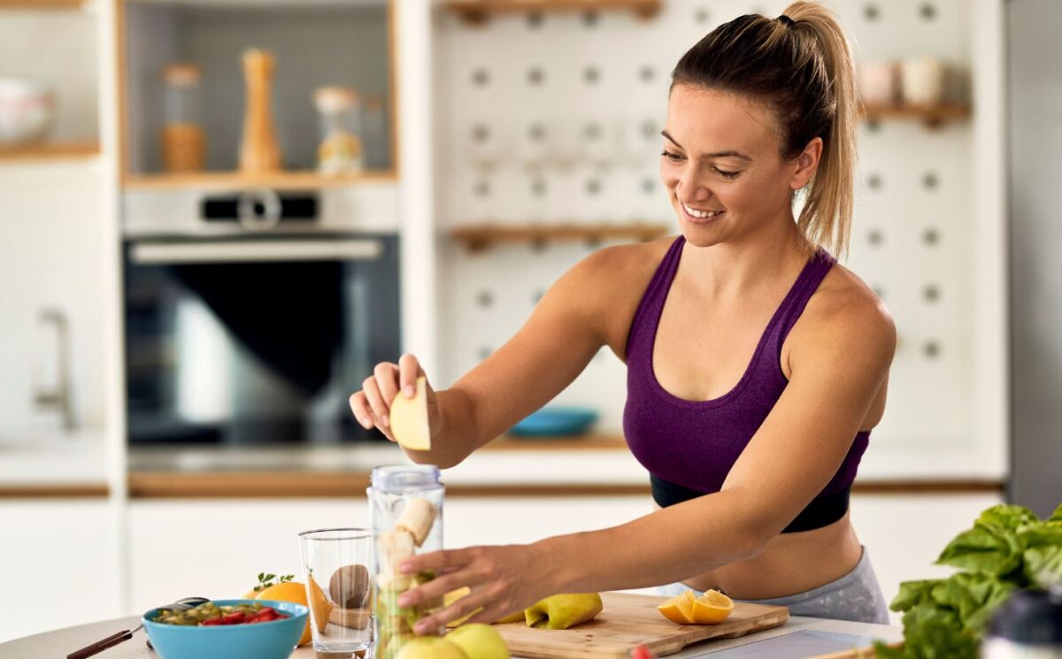 Sporty woman making a fruit smoothie on a kitchen island, to illustrate an article about meal timing health benefits.