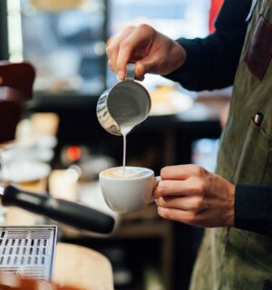 Photo shows a barista in a coffee shop or café pouring foamed milk into a cup of coffee. According to a new survey, the majority of UK coffee shops are open to making plant-based milk the default option