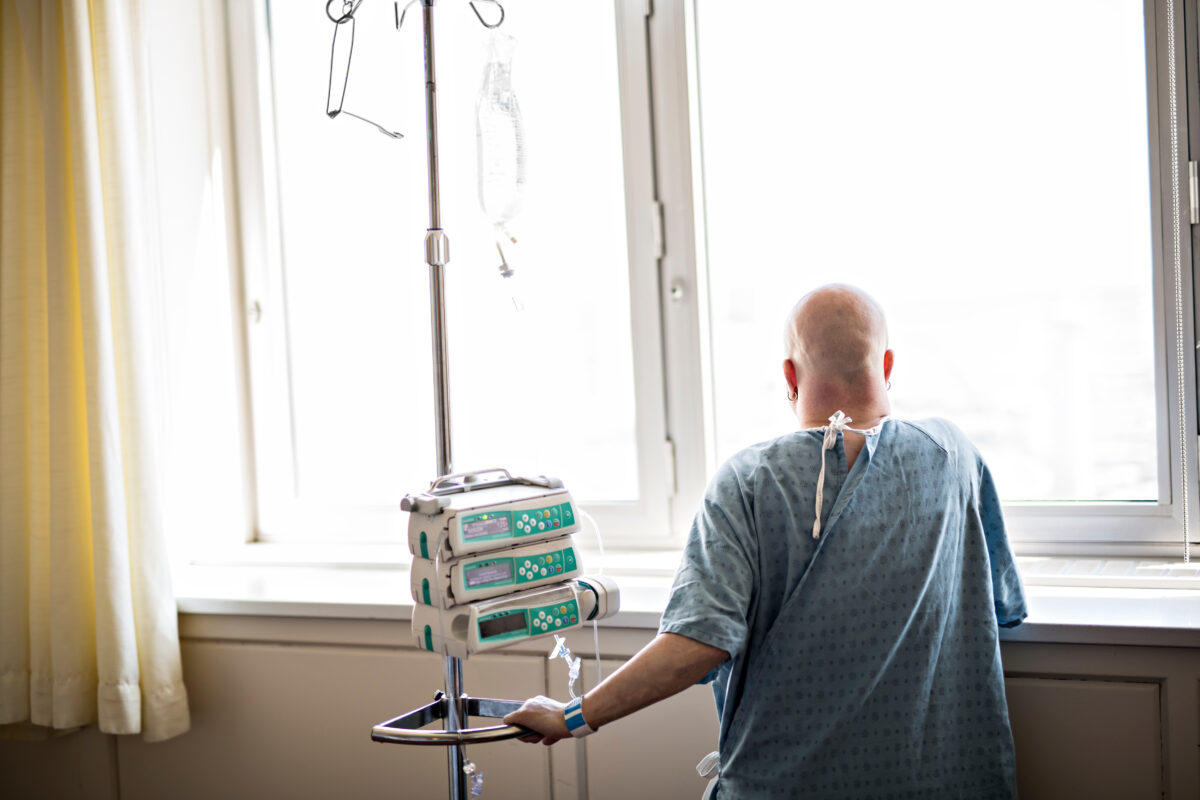 Photo shows a woman standing at a hospital window with an IV stand next to her