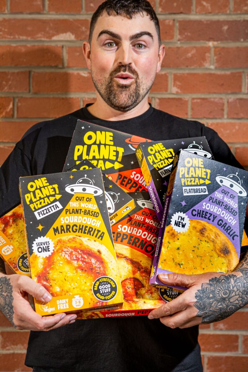 Photo shows Joe Hill, the co-founder of One Planet Pizza, holding a selection of the brand's frozen vegan pizzas and cheesy garlic bread. One Planet Pizza has passed £1 million in annualised retail sales for the first time ever