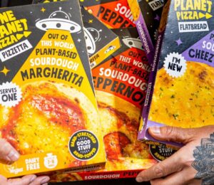 Photo shows Joe Hill, the co-founder of One Planet Pizza, holding a selection of the brand's frozen vegan pizzas and cheesy garlic bread. One Planet Pizza has passed £1 million in annualised retail sales for the first time ever