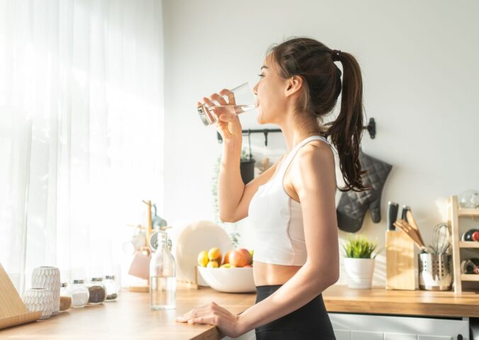 Woman in sportswear drinking water in the kitchen, showing that hydration is one of 8 daily habits to reduce inflammation