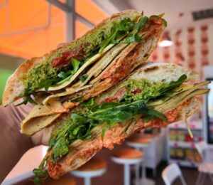 Reyes holding a sandwich at Maciel's Plant-Based Butcher Shop with the deli in the background, to illustrate an article about the award-winning vegan sandwiches in LA