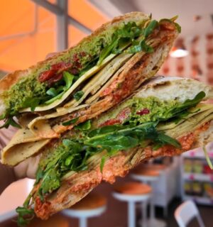 Reyes holding a sandwich at Maciel's Plant-Based Butcher Shop with the deli in the background, to illustrate an article about the award-winning vegan sandwiches in LA