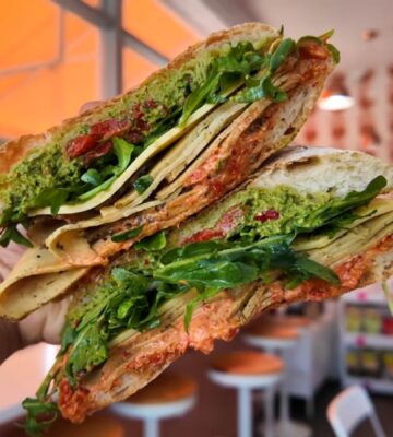 Reyes holding a sandwich at Maciel's Plant-Based Butcher Shop with the deli in the background, to illustrate an article about the award-winning vegan sandwiches in LA