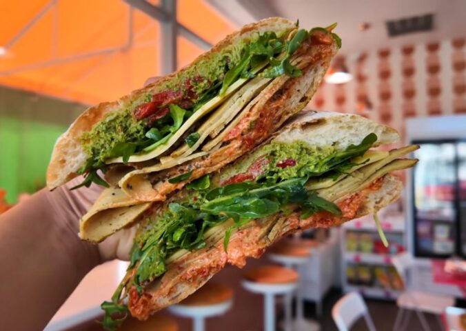 Reyes holding a sandwich at Maciel's Plant-Based Butcher Shop with the deli in the background, to illustrate an article about the award-winning vegan sandwiches in LA