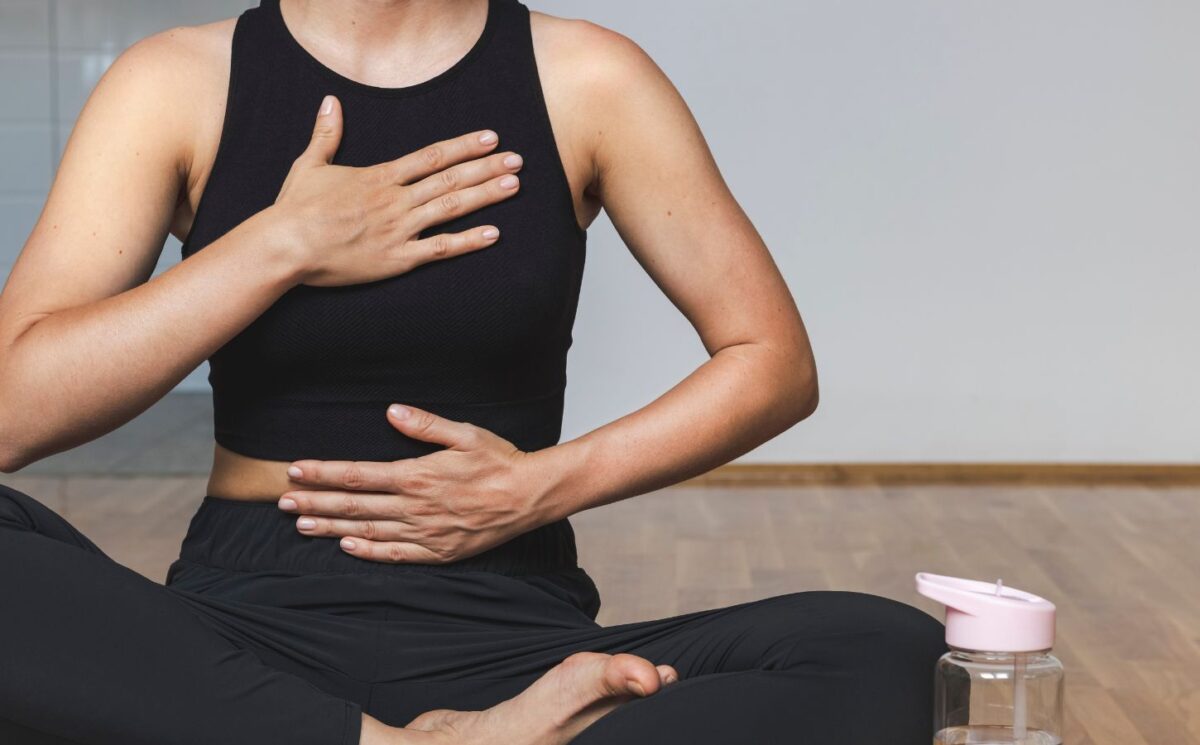 A woman in yoga clothes sits in the lotus position with one hand on her belly and the other on her chest while practicing diaphragmatic breathing, illustrating how to breathe better.