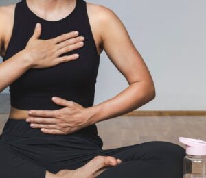 A woman in yoga clothes sits in the lotus position with one hand on her belly and the other on her chest while practicing diaphragmatic breathing, illustrating how to breathe better.