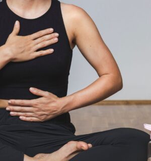 A woman in yoga clothes sits in the lotus position with one hand on her belly and the other on her chest while practicing diaphragmatic breathing, illustrating how to breathe better.
