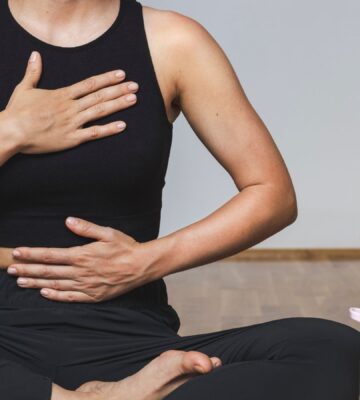 A woman in yoga clothes sits in the lotus position with one hand on her belly and the other on her chest while practicing diaphragmatic breathing, illustrating how to breathe better.