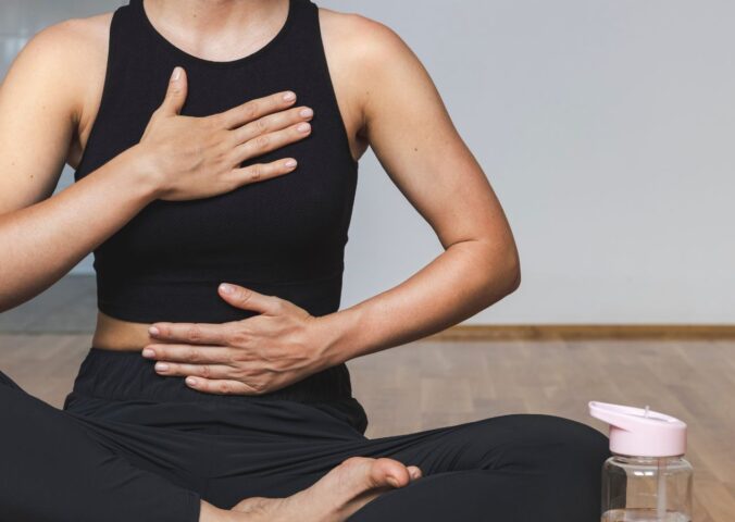 A woman in yoga clothes sits in the lotus position with one hand on her belly and the other on her chest while practicing diaphragmatic breathing, illustrating how to breathe better.