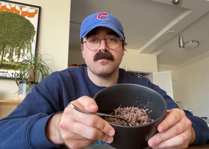 During Eddy Burback's carnivore diet experiment he sits at a table in his apartment holding a bowl of ground beef toward the camera