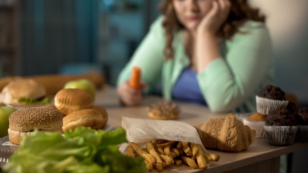 A depressed-looking woman sits at a table surrounded by junk food while holding a carrot, to illustrate an article about stress eating and weight loss
