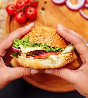 Photo shows a woman's hands holding a mushroom burger with a wooden chopping board and ingredients in the background. A new study has found that generative AI can help create more delicious, sustainable, and nutritious burgers, including plant-based options.