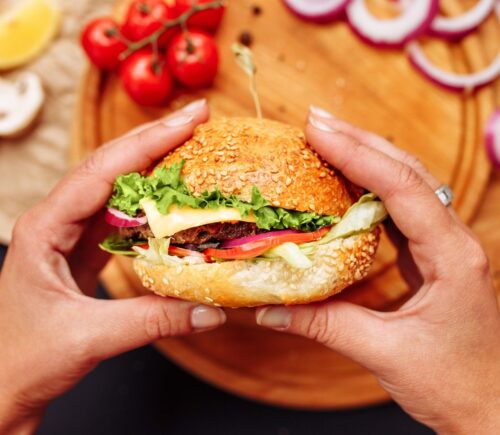 Photo shows a woman's hands holding a mushroom burger with a wooden chopping board and ingredients in the background. A new study has found that generative AI can help create more delicious, sustainable, and nutritious burgers, including plant-based options.
