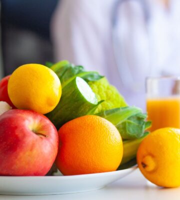 Photo shows a large bowl of fresh fruit and vegetables on a table, as someone in a lab coat writes on a clip board. Dr Mathew Nagra has debunked claims that Bryan Johnson’s recent diagnosis of autoimmune gastritis (AIG) was caused by a vegan diet