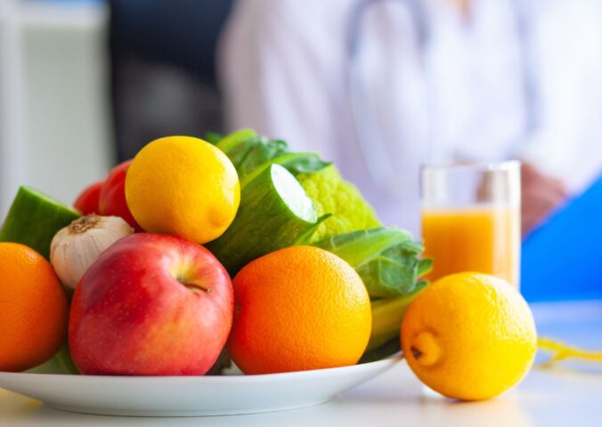 Photo shows a large bowl of fresh fruit and vegetables on a table, as someone in a lab coat writes on a clip board. Dr Mathew Nagra has debunked claims that Bryan Johnson’s recent diagnosis of autoimmune gastritis (AIG) was caused by a vegan diet